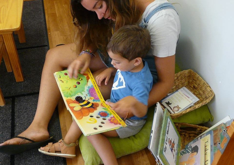 Rockingham Montessori School infant toddler seated on the floor with her sister reading him a book