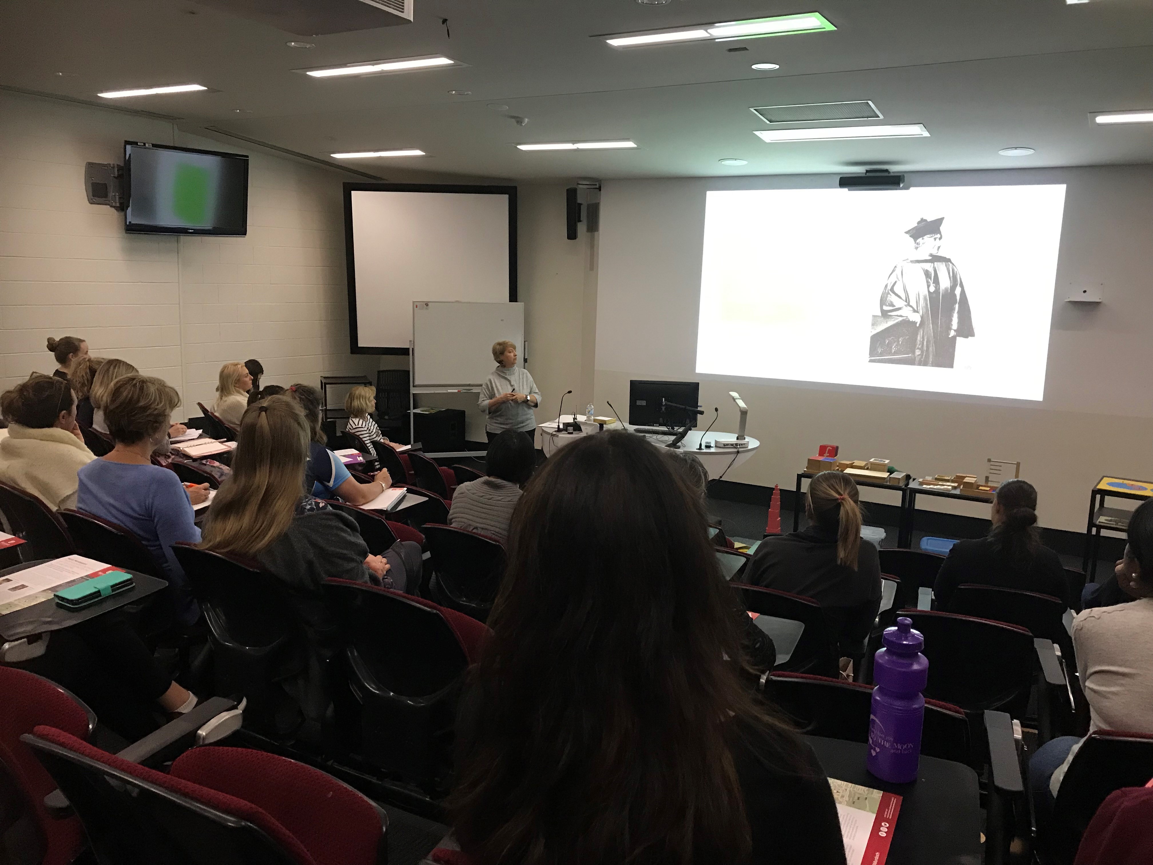 Rockingham Montessori male and female students listening to the instructor in front of the classroom as they discuss something on the projected screen