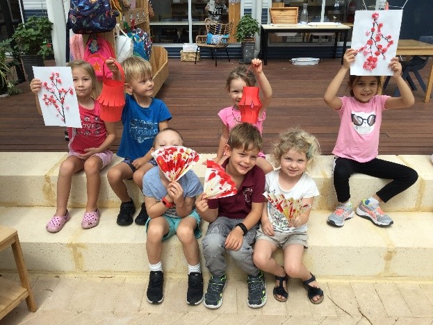 Rockingham Montessori students holding their paper flower artwork while seated on the ground outside their classroom